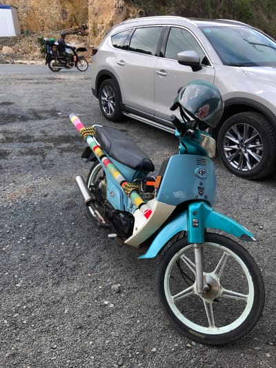 In Nam Hải, Vietnam, a light-colored SUV is parked on a gravel lot. In front of the SUV, a light blue moped is parked. The moped has a teal-colored body and a black seat. A green helmet with a red emblem is placed on the handlebars. The rear rack of the moped is decorated with colorful yarn wrapping. A second motorcycle, ridden by a person wearing a helmet and camouflage pants, is visible in the background, loaded with bundles of greenery. The environment consists of a rocky hillside and a paved road.