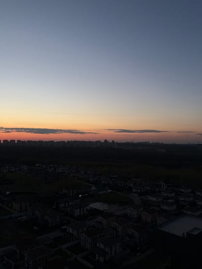 The image shows an aerial view of Анкудиновка, Russia at sunset or sunrise. The sky transitions from blue to orange. A line of clouds is visible, and beyond it, the skyline of a distant cityscape. The foreground shows residential buildings and green space. The buildings appear dark and silhouetted.