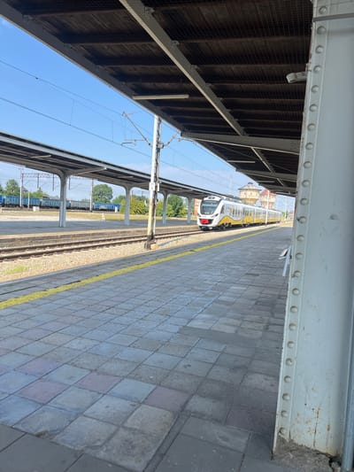A train with yellow and white stripes is stopped at a platform in Kamieniec Ząbkowicki, Poland. The train is on the tracks, and there is a yellow line on the platform. The platform is made of concrete tiles. There is a covered walkway over the platform, supported by columns. The sky is blue. In the background, there are trees and buildings, including a water tower. The image is taken from a low angle, looking down the platform.