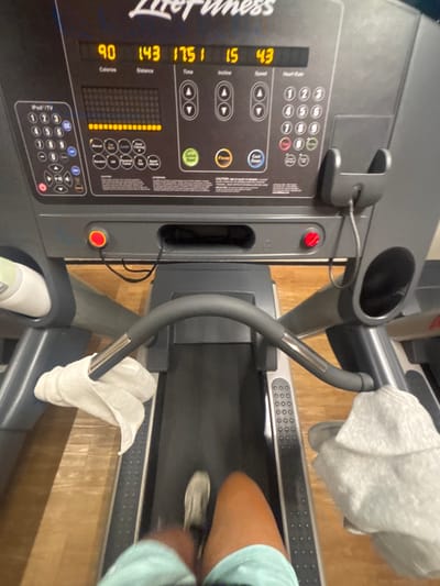 This media file captures an overhead view of a person exercising on a Life Fitness treadmill in an indoor setting, likely a gym or home gym, in Komchén, Mexico. The image is rotated counter-clockwise, showing the treadmill's console on the left and the running belt extending to the right.
The central subject is a gray Life Fitness treadmill. Its console, prominent in the left half of the frame, displays various workout metrics. Visible readouts include "Calories: 90", "Distance: 143", "Time: 17:51" (indicating 17 minutes and 51 seconds), "Incline: 15", and "Speed: 4.3" (likely mph or kph). Below these, a horizontal bar graph of illuminated yellow segments suggests an intensity or progress indicator. The console features a numeric keypad (0-9), arrow buttons for "Speed" and "Incline" adjustments, a "Quick Start" button, a "Pause" button, and controls labeled "iPod/TV". A smartphone holder with a cable connected is positioned on the top left of the console, and a red emergency stop button is clearly visible to the right of the main display area. The brand name "Life Fitness" is partially visible along the left edge of the console.
On the treadmill belt, the lower legs and feet of a person are visible, wearing light blue shorts and white or light-colored athletic shoes. The slight motion blur on the legs and shoes indicates that the person is actively running or walking. A white towel is draped over the lower left handlebar, and another gray towel rests on the upper right handlebar. A white water bottle is placed in a holder on the right side of the treadmill. The floor beneath and surrounding the treadmill is a light-colored wood or laminate. The scene conveys an active workout session in a fitness environment.