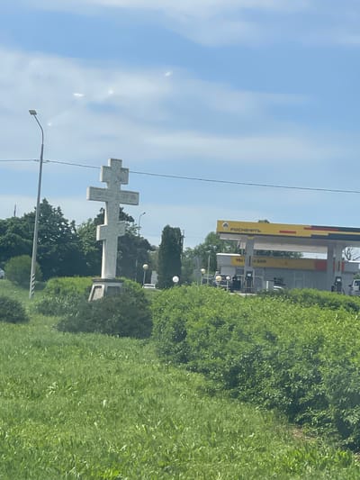 A roadside scene depicts a large white Orthodox cross erected as a monument in Donskoe, Russia, set against a backdrop of trees and a clear, blue sky. Overhead power lines and a street lamp are visible near the cross. In the distance, a "Rosneft" gas station stands at the roadside, adjacent to dense greenery.