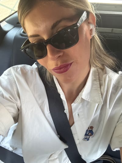 A close-up, selfie-style shot shows a blonde woman with dark sunglasses and lipstick, wearing a white button-down shirt with a polo bear patch. She appears to be in the back seat of a vehicle, as indicated by the dark interior and seatbelt. The background is blurred, suggesting the car is in motion. It is not possible to determine the specific location within Nanterre, France from the image.