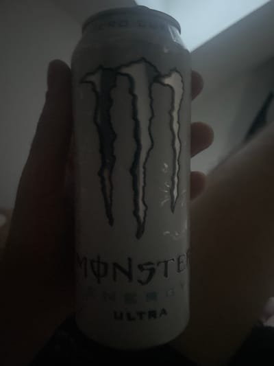 A person's hand is holding a white can of Monster Energy drink. The can has the Monster Energy logo printed on it, which consists of three claw marks. Below the logo, the word "MONSTER" is visible, followed by "ENERGY" and "ULTRA". The background is dark and blurry, with some hints of light in the upper right corner, suggesting an indoor environment. The image quality is poor due to low light conditions. There is no information to suggest the location is in Paczkowo, Poland.