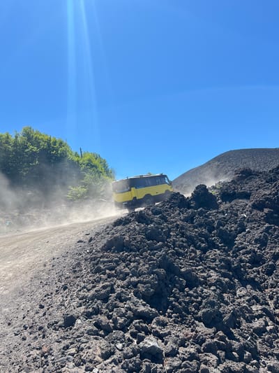 A yellow tour van drives on a dirt road through a rocky, volcanic landscape under a clear blue sky. The van is kicking up dust as it moves. The terrain is uneven, with large, dark rocks and sparse green vegetation visible on the left side of the frame. The overall scene suggests an off-road excursion in a volcanic area, likely Mount Etna, given the location in Linguaglossa, Italy.