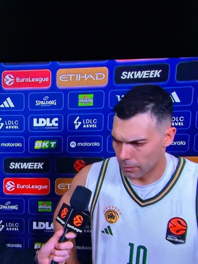 A basketball player in a white and green jersey labeled '10' stands in front of a sponsor board during a EuroLeague interview. The board displays various brand logos, including EuroLeague, Etihad, Spalding, Uludag Gazoz, LDLC ASVEL, Motorola, BKT, and SKWEEK. The player looks downwards while holding a microphone. He wears the Adidas logo on his jersey.
