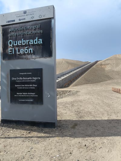 The image depicts a large sign in front of a landscape with a long staircase leading up a hill. The sign, constructed of stone and featuring a dark plaque, indicates that an "Integral Solution against Floods" has been implemented in "Quebrada El León," a ravine in the city of Huanchaco, Peru. It lists the inaugurating officials as Dina Ercilia Boluarte Zegarra, Gustavo Lino Adrianzén Olaya, and Hernán Yaipén Aréstegui, and states that the inauguration took place in La Libertad in January of 2025. The landscape behind the sign is arid, with a sandy ground and a hill that has been landscaped with a concrete staircase for access. A wooden fence is visible to the right. The sky is partially cloudy.