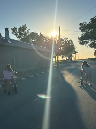 Two young girls are seen on a road in Enevo, Bulgaria, at dusk. The girl on the left, closer to a building and with her back to the camera, is pushing a bicycle. She has long, brown hair and is wearing a light-colored shirt and shorts. The girl on the right is riding a bicycle with training wheels. She also has long, brown hair and is wearing a light-colored shirt and shorts. The sun is setting, casting long shadows across the asphalt road. Trees and power lines are visible in the background, along with a building that appears to be a barn or shed. The sky is a clear blue with a faint haze from the setting sun.