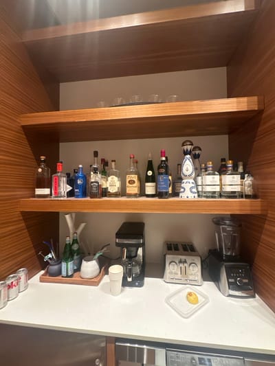 The image depicts an interior space, specifically a well-appointed bar or kitchen area, consistent with a residence or hotel located in Aspen, United States. The scene is illuminated by internal lighting, showcasing a modern design with prominent wooden shelving and paneling. No individuals are present, and no actions or interactions are occurring.
The setup includes a white countertop with two floating wooden shelves above it. The uppermost visible shelf holds a dark tray with four clear small glasses and two additional clear glasses to its right. The middle shelf displays approximately 16 bottles of various alcoholic beverages, including clear spirits (e.g., Hendrick's Gin, Tito's Vodka, LALO Tequila), dark spirits (e.g., Jack Daniel's Whiskey), and wine. A distinctive white and blue patterned ceramic bottle, likely tequila, stands centrally on this shelf.
On the white countertop, from left to right, are three silver beverage cans (one identifiable as Coca-Cola), a dark ceramic holder containing stirrers/straws, two green glass bottles of sparkling water, and a white ceramic vessel with additional stirrers/straws, all arranged on a woven tray. Further right, a stack of white paper cups, a black coffee maker, a silver four-slice toaster, and a large black and silver blender are visible. A clear plastic container with a white lid holds a single yellow, crescent-shaped food item. The area beneath the countertop features stainless steel appliance fronts or drawers.
