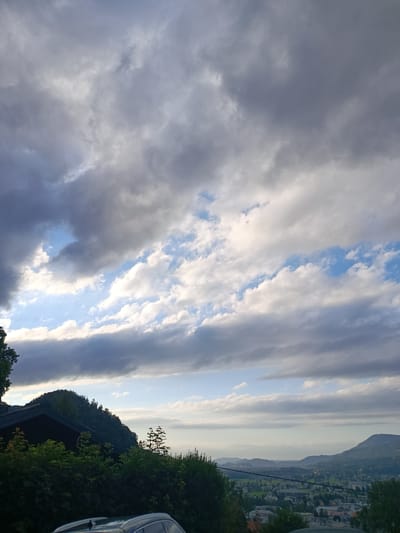 The image shows a panoramic view of Hallein, Austria, dominated by a dramatic sky filled with heavy, dark clouds interspersed with patches of bright blue sky and wispy white clouds. In the foreground, a dark-colored vehicle is partially visible at the bottom edge. To the left, a dark-roofed building with a dark facade sits on a steep, tree-covered hillside, with dense green foliage obscuring much of the lower portion of the structure. Further down the hillside, a town or city sprawls across the landscape, with various buildings and infrastructure visible. Rolling hills and mountains form the backdrop, fading into the distance under the cloudy sky. The lighting suggests it is late afternoon or early evening, with the sun casting a soft glow on the clouds and landscape.