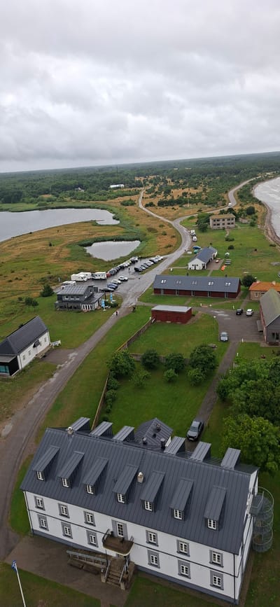 This aerial photograph depicts a coastal landscape in Sääre, Estonia, under an overcast sky. The scene features a body of water, possibly a bay or inlet, on the left, with a shoreline characterized by marshy vegetation and scattered trees. A road winds through the landscape, connecting various structures.

Prominent among the structures is a large, modern building with a dark gray, multi-pitched roof and white siding, featuring numerous windows. To the right of this building, a narrow, light-colored staircase with railings leads up to a platform. Further to the right, two large, circular metallic structures, resembling wind turbines or observation decks, are visible on top of another section of the building.

Scattered throughout the scene are smaller buildings, including red, rectangular structures and other modern-looking residences or apartments with dark roofs. A parking lot filled with several cars and what appear to be small boats or kayaks is situated along the road near some of these buildings. A few individuals can be seen scattered among the vehicles and buildings, suggesting activity and perhaps a sense of community or a public space. The overall impression is one of a developed coastal area with a blend of residential and potentially recreational facilities. The weather appears to be cloudy and potentially humid, typical of a coastal environment. No visible text is present in the image.