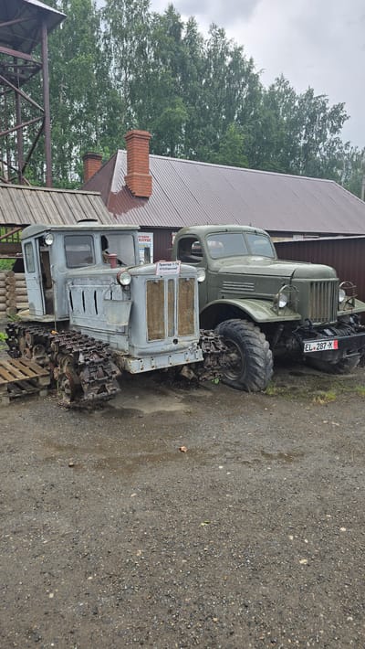 The image shows a dusty, outdoor scene with two vintage vehicles parked on gravel. In the foreground, a gray tracked tractor, possibly a DT-74 based on the sign, is positioned left of center. It has visible rust on its tracks and a bucket hanging from its front. To the right of the tractor, a green military-style truck is parked. The truck has a license plate that reads "EL 287 X". Behind the vehicles, a brown-roofed building with brick chimneys is visible, along with a metal tower structure to the far left. The background consists of a dense line of green trees under a cloudy sky. The environment appears to be a museum or an exhibition of historical machinery. The location is stated to be in the city of Bol'shoi Mashlyak, Russia. There are no people visible in the image.