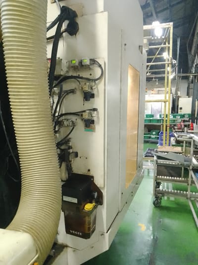 The image depicts a close-up view of industrial machinery, likely in a factory setting. A large, ribbed, beige hose is prominently featured on the left side of the frame, curving upwards. To its right, the side of a white industrial machine is visible, featuring various components including electrical conduits, pneumatic valves, and two small digital displays showing "0.12". A black, rectangular container with a yellow cap is attached to the lower part of the machine, with "VOGEL" visible on it. In the background, a yellow metal structure with multiple horizontal bars can be seen, along with shelving units holding green containers and what appears to be a conveyor belt system with spherical objects. The floor is a shiny green, with some water or liquid present. The overall scene suggests an operational or recently used manufacturing environment. This image does not provide any specific geographical context that would allow for pinpointing a location within the city of Ciantra, Indonesia.