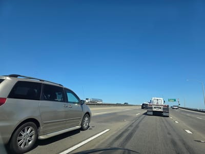 The media file captures a view from a vehicle traveling on a multi-lane highway under a clear blue sky, suggesting a daytime setting with good weather. In the foreground, the rear of a gold minivan is visible on the left, and the hood of another vehicle from which the photo is taken is at the bottom. The highway is well-lit by the sun, and lane markings are clearly visible on the asphalt.
Several other vehicles are present on the highway, including a flatbed truck with the number "4529" on its rear, a white truck with a trailer, a black SUV, and other cars. These vehicles suggest ongoing traffic flow on the highway.
In the background, a sign for "Jefferson Blvd" and "Stockton Blvd" is visible, along with an exit ramp. This indicates the location is in Sacramento, United States of America. The overall scene conveys a sense of everyday travel on a busy road. There are no visible people, and the emotions are neutral, reflecting a standard highway environment.