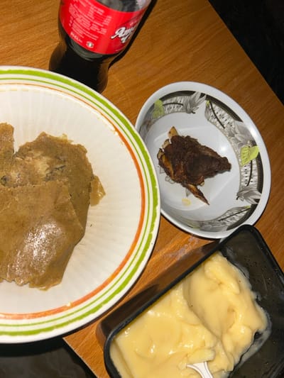 The image displays a meal arranged on a wooden table, likely in an indoor setting in Debasco, Nigeria. In the upper left, a large portion of light brown, dense food, consistent with a swallow food like Amala or Eba, is contained within a white ceramic bowl featuring green and orange stripes on its rim. To the right of this bowl, a smaller white ceramic bowl, decorated with a grey and black leaf pattern, holds several pieces of dark brown, roasted meat, possibly ribs. At the top of the frame, a dark beverage in a clear plastic bottle with a prominent red label, partially visible as "Amigo" and indicating "35 CL," rests on the table. In the lower right corner, a black rectangular plastic container holds a pale yellow, semi-liquid substance, resembling custard or pap, with a small white plastic spoon inserted into it. No individuals are visible.