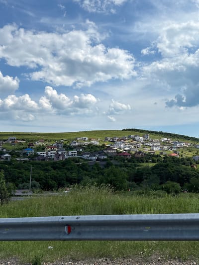 This image depicts a residential area situated on verdant, rolling hills in Varvarovka, Russia, under a partially cloudy blue sky during daylight hours. In the immediate foreground, a silver metal guardrail extends horizontally across the bottom of the frame, featuring a red and white reflector mounted near its center. Beneath and behind the guardrail, coarse gravel and tall green grass are visible. A dense band of dark green trees occupies the lower midground, separating the foreground from the cluster of houses. Numerous single and two-story houses are visible across the hillsides, showcasing varied architectural styles, some with white facades and modern designs, others with traditional colored roofs (red, blue, brown). The houses are interspersed with patches of green grass and foliage. The hills gently slope upwards, culminating in a relatively flat, green horizon line under a sky dominated by scattered, puffy white cumulus clouds with some lighter cirrus streaks. No individuals, specific actions, or interactions are discernible within the frame.
