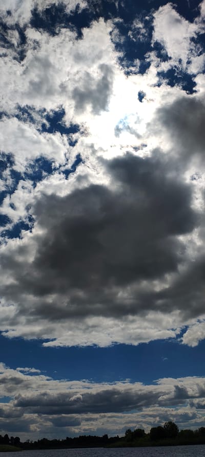 The image shows a sky filled with dramatic clouds over a body of water. The sky transitions from a deep blue at the bottom to brighter areas where the sun is partially obscured by thick, dark gray clouds. There are also patches of white, wispy clouds scattered throughout. The lower portion of the image reveals a dark, silhouetted treeline along the horizon, separating the sky from the calm water. The scene appears to be a landscape, possibly near Щиглицы, Russia, given the information provided. No people or specific objects are visible other than the natural elements of sky, water, and vegetation. The lighting suggests it is daytime, with the sun's rays creating a contrast between the darker and lighter clouds.