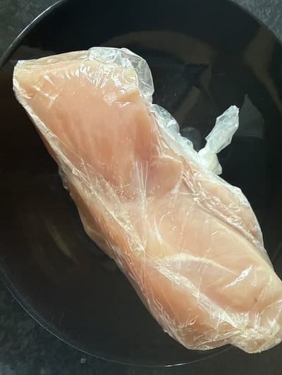 This is a close-up, overhead shot of a raw chicken breast wrapped in clear plastic wrap. The chicken breast is the primary subject, positioned diagonally in the center of the frame. It is light pinkish-white in color and appears to be a substantial cut. The plastic wrap is crinkled and somewhat transparent, allowing the texture and form of the chicken to be visible. There are some slight reflections on the plastic.

The chicken is resting on a dark, circular plate, likely black or very dark gray. The plate has a subtle sheen, reflecting some light. The background behind the plate is dark and out of focus, suggesting an indoor setting, possibly a kitchen counter.

No people are visible in the image, nor is there any text. The lighting appears to be even and overhead, with no strong shadows or indications of a specific time of day or weather conditions. The overall impression is one of food preparation or storage. The context provided indicates the location is Drogheda, Ireland, but there are no visual cues within the image itself to confirm this.