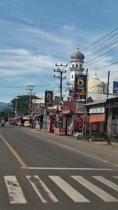 The image depicts a street scene in Teungoh Baro, Indonesia, on a bright, partly cloudy day. A white mosque with a grey dome and minaret is visible in the background, along with a second mosque with a golden dome. Several power lines crisscross the sky. In the foreground, a two-lane asphalt road runs from left to right, with a pedestrian crossing marked by white stripes. On the right side of the road, a row of small businesses and food stalls line the street. Some stalls have awnings and advertising banners, including one for "DIARUM 15.000". On the left side of the road, a few motorcycles are seen, with one rider wearing a helmet. The overall impression is of a bustling local street with religious buildings and commercial activity.