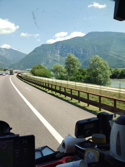 The image captures a view from inside a vehicle, likely a truck or RV, looking forward through the windshield. The vehicle is traveling on a multi-lane highway with a white line marking the lanes. The highway curves to the left, bordered by a metal guardrail and green vegetation. Beyond the guardrail, a river is visible, and further in the distance, lush green mountains with rocky outcrops rise against a clear blue sky with scattered white clouds. The mountains appear to be in the vicinity of Volano, Italy, as suggested by the terrain and foliage.

In the foreground, inside the vehicle, several objects are visible on the dashboard. A GPS device or navigation unit with a screen displaying "80" within a red circle (likely a speed limit indicator) and other digital information is prominent on the left. To the right, other items include a roll of toilet paper, a yellow mug, a black electronic device with a green light, and a white electric kettle. The dashboard also shows some smudges or dirt on the windshield.

The overall scene depicts a journey through a scenic mountainous region, likely on a sunny day, with the driver's perspective focused on the road ahead and the surrounding natural landscape.