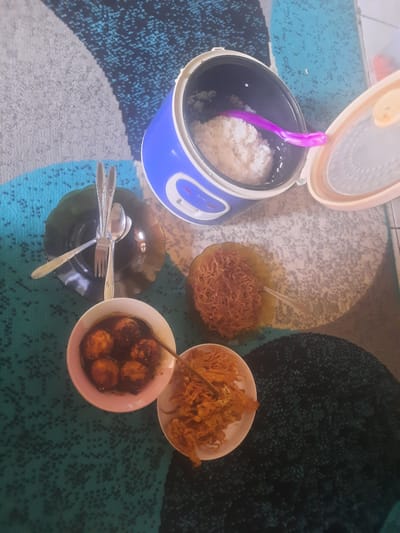 This is a high-angle, medium shot of a meal laid out on a patterned rug. In the upper right is an open blue and white rice cooker filled with white rice, with a purple spoon resting inside. To the left of the rice cooker, there is a dark green, fluted plate with a fork and two spoons resting on it. Below this, to the left, is a small white bowl containing several brown, spherical items in a dark sauce. To the right of that bowl is another white bowl filled with crispy, thin, fried noodles. Further to the right, and slightly lower, is a third white bowl containing more of the crispy noodles. The rug has a textured pattern of light grey, dark grey, and turquoise.