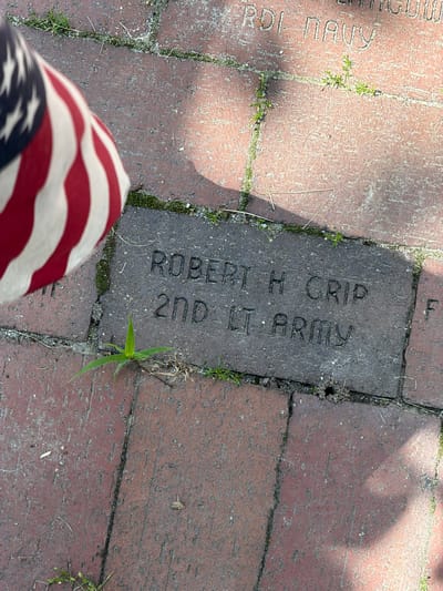 The image shows a portion of an American flag and an engraved brick on a brick walkway. The brick is engraved with the name "ROBERT H CRIP" on the top line and "2ND LT ARMY" on the bottom line. The flag is red, white, and blue. The brick walkway is made of red bricks and is partially covered with grass and dirt. The scene appears to be outdoors. There is no indication that this scene is located in Peterborough, United States of America.