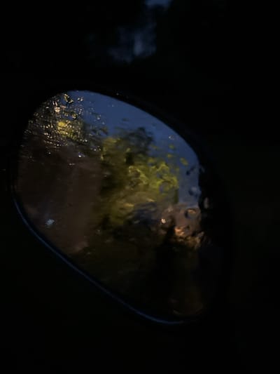 The image shows a close-up view of a car's side mirror at night. The mirror is wet with raindrops, distorting the reflection of the surroundings. The reflection shows blurred shapes of trees with yellow and green foliage, likely illuminated by streetlights or house lights. There are also some faint lights that could be from buildings or vehicles. The overall scene is dark and moody, suggesting a rainy evening in Sergeevka, Russia. No people or specific actions are discernible in the image due to the low light conditions and the nature of the reflection.