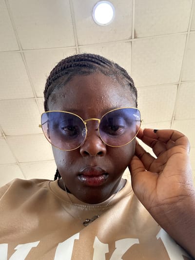 The image shows a dark-skinned person with braided hair, wearing sunglasses with gold frames and blue lenses. Their right hand is touching the top of the glasses frame. They are wearing a tan and white shirt and a silver necklace. The background appears to be a tiled ceiling with a light fixture. There is no indication of this image being in the city of Madangala, Nigeria.