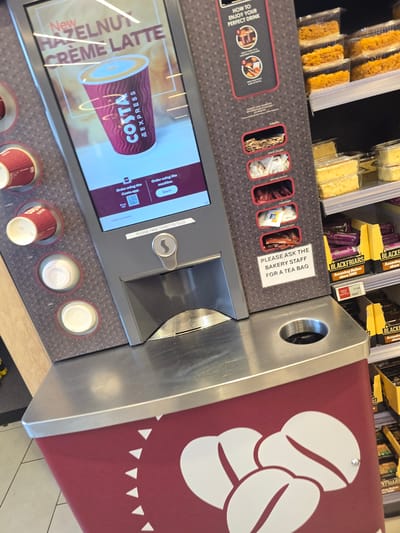 The image shows a Costa Express coffee machine with a digital display. The display advertises a "New Hazelnut Crème Latte" and shows a cup of coffee with the Costa Express logo. Below the display, there are slots for various additions to the drink, including stirrers, white sugar, brown sugar, sweetener, and chocolate dust. A sign on the machine reads "PLEASE ASK THE BAKERY STAFF FOR A TEA BAG". In the foreground, there is a red base with a white coffee bean design. The machine is situated next to shelves stocked with packaged cakes and confectionery, specifically "BLACKFRIARS" branded items. The overall environment appears to be a retail setting, possibly a supermarket or convenience store. The image does not provide specific details about the city of Stockton-on-Tees, United Kingdom.