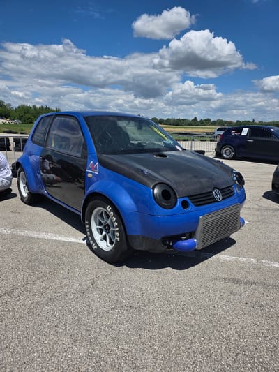 A modified blue Volkswagen Lupo is parked on an asphalt surface, likely a racetrack or parking lot, under a bright blue sky with scattered white clouds. The car features a carbon fiber hood with prominent latches and circular openings, a large front-mounted intercooler, and wide fender flares. The wheels are white with racing tires. A person in white shorts is partially visible kneeling to the left of the car. In the background, other vehicles and barriers are present, along with a tree line and distant fields. The scene appears to be a sunny day with good visibility. There is no information within the image to place it specifically in Akatsievo, Bulgaria.