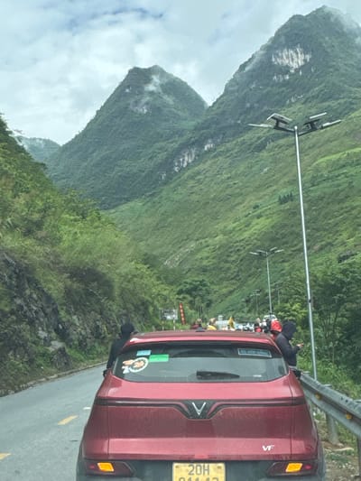 The image shows a red VinFast VF e34 car with the license plate 20H-041.13 parked on the side of a road, likely taken from another vehicle. The background features steep, green, cloud-covered mountains. The scene appears to be in a mountainous region, and according to the license plate number and general scenery, the location is most likely in the Yên Minh Commune, Vietnam. A crowd of people can be seen standing further up the road beyond the car. There is a guardrail along the side of the road, and streetlights extending into the scene.