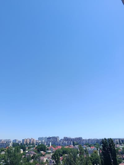 This image captures a panoramic view of the residential areas of Odesa, Ukraine, on a clear day. The foreground is dominated by a dense canopy of lush green trees, interspersed with houses featuring red tile roofs and lighter-colored walls. Beyond these lower-rise dwellings, a skyline of mid-rise and high-rise apartment buildings stretches across the horizon, characteristic of urban development. The sky is a uniform, vibrant blue, devoid of clouds, suggesting fair weather. The overall scene depicts a serene, lived-in urban landscape with a blend of residential housing and taller apartment complexes, typical of a city like Odesa.