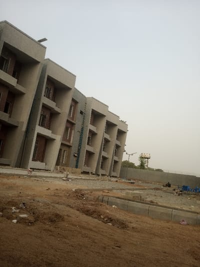 The scene depicts an outdoor area in Bwari, Nigeria, featuring a multi-story building and undeveloped ground. A large building, positioned diagonally across the left and center, presents a light grey, angular concrete facade with recessed sections and projecting ledges or balconies across its visible three to four stories. Dark rectangular openings indicate windows or unglazed frames. A vertical pipe or conduit system is visible on one section of the building. The foreground consists of disturbed, reddish-brown earth with scattered debris, including plastic bags, and sections of uncompleted concrete curbing and gravel. To the right, a grey concrete wall topped with barbed wire runs horizontally. Two individuals are seated on the ground near this wall, dressed in dark clothing, with several blue plastic chairs grouped nearby. In the background, beyond the wall, a tall metal tower with a white rectangular structure at its apex stands against a uniformly light grey, hazy sky.