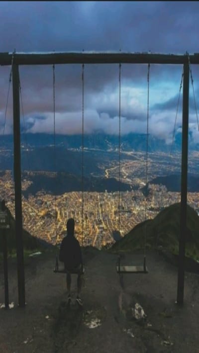 An individual sits on a swing overlooking a cityscape illuminated at night. The swing set, crafted from dark wood, stands atop a hill with greenery visible. Above, clouds obscure the sky, creating a twilight ambiance. A second swing hangs empty nearby. The city lights stretch across the valley, contrasting with the dark silhouette of the person on the swing.