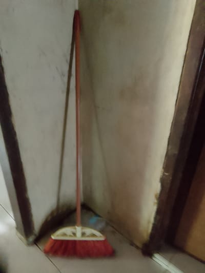 A red-bristled broom with a light-colored handle rests against a light-colored, textured wall in a corner. The broom's handle is long and slender, and it leans at an angle. The bristles are densely packed and a vibrant red. The wall appears to be a neutral tone, possibly off-white or beige, with some subtle discoloration or smudges. The scene is dimly lit, creating soft shadows. The broom is positioned in what appears to be an interior space, possibly a home or a utility area. There is no indication of the city of Merbopuntong, Indonesia, in this image.