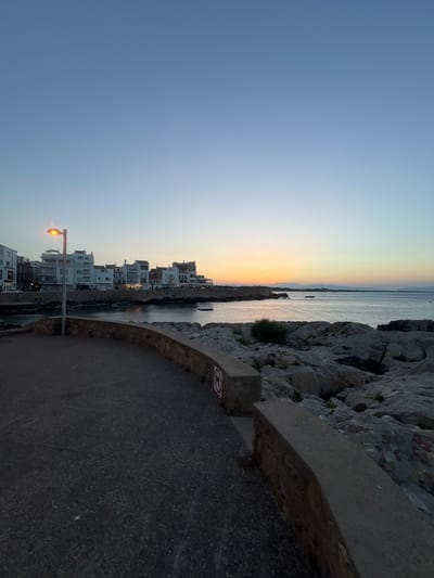 The image depicts a coastal scene in L'escala, Spain, during twilight. A paved walkway curves along the foreground, leading the eye towards a body of water. On the left, a streetlamp casts a warm glow over a section of the walkway. Beyond the water, a line of white buildings stretches across the horizon, indicative of a town or village. The sky transitions from a deep blue at the top to lighter hues of orange and yellow near the horizon, where the sun has recently set. Rocky terrain lines the right side of the image, contrasting with the smoother man-made structures and walkway. The sea is calm, with a few small boats visible in the distance. A circular sign with a red border and a black symbol is affixed to a low wall in the middle right of the frame. The overall atmosphere is peaceful and serene, characteristic of early evening in a Mediterranean coastal town.