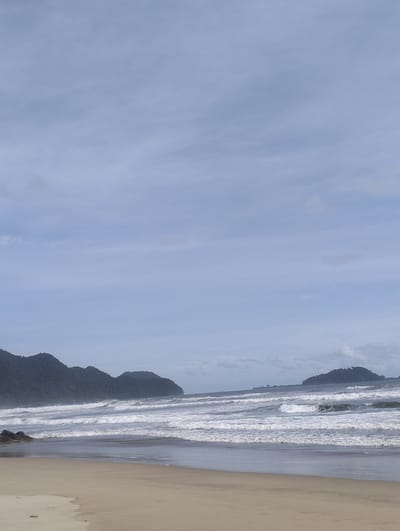 A wide, sandy beach stretches across the foreground of this image, with the gentle, tan-colored sand meeting the calmer waters at the shoreline. Several rows of white-capped waves are breaking and rolling towards the beach, indicating a moderate surf. In the middle ground, the ocean extends outwards, with distinct lines of white foam marking the successive waves. In the background, a chain of dark green, heavily forested mountains forms a dramatic backdrop to the scene. To the right, a smaller, solitary island covered in dense foliage rises from the sea, also appearing mountainous. The sky above is a soft, pale blue, with scattered, wispy clouds that create a slightly overcast but bright atmosphere. The overall impression is of a natural, undeveloped coastline with a serene, natural beauty. This scene is located in Meudheun, Indonesia.