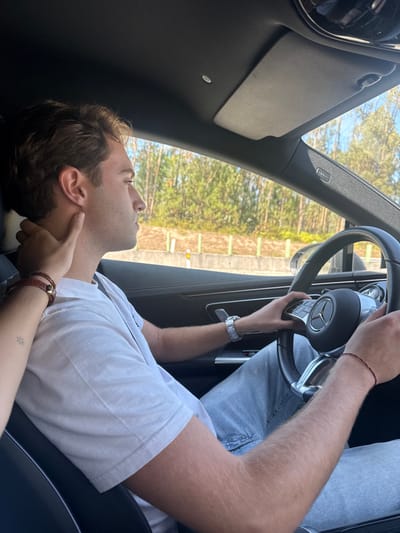A daylight, medium shot captures the interior of a vehicle, depicting a male driver and a female's hand. The male, seated in the driver's position, has light brown hair, wears a white short-sleeved t-shirt, light blue jeans, a silver-toned wristwatch on his left wrist, and a thin red bracelet on his right. His hands grip a black multi-functional steering wheel with a Mercedes-Benz emblem. His head is turned slightly to the right, and his gaze is directed forward. A female's left hand, adorned with a small star tattoo on the forearm and two brown leather bracelets, rests on the male driver's neck and jawline from the left side of the frame. The vehicle's interior features dark-colored headliner and door panels, a light gray sun visor, and a visible "AIRBAG" label on the A-pillar. Through the car windows, a bright, clear sky is visible above a dense backdrop of green evergreen trees, indicating a forested area, with a white and yellow roadside barrier separating the road from the vegetation. The scene is depicted as taking place on a road in the city of Pombal, Portugal.