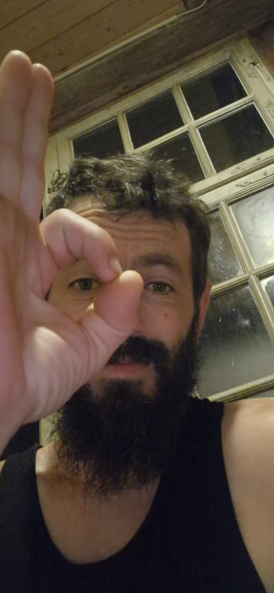 The image, taken indoors, features a close-up of a man's face. He has dark, curly hair, a full beard, and brown eyes. His left hand is held up, with his thumb and index finger forming a circle, positioned near his right eye, partially obscuring his face. He is wearing a dark, sleeveless shirt. In the background, a window with multiple panes and a wooden ceiling are visible. The context provided about Wattenwil, Switzerland, is not directly observable in the image itself.