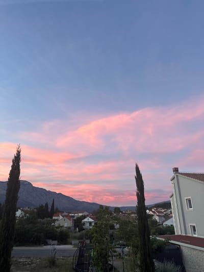 The image depicts a tranquil sunset scene in Grad Korčula, Croatia. The sky is a gradient of soft blues and vibrant pinks, painted with wispy clouds. In the foreground, two tall, slender cypress trees stand sentinel. Beyond them, a cluster of houses with red-tiled roofs are nestled amongst lush greenery. In the distance, a majestic mountain range forms a dramatic backdrop. The overall atmosphere is one of peaceful serenity, typical of a Mediterranean evening. There are no people or animals visible in the image. The scene is composed of natural elements and man-made structures, contributing to a picturesque view.