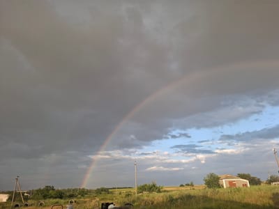 This is a wide-angle, medium-shot, eye-level photograph taken in Chorna, Ukraine. The scene depicts a rural landscape with a dramatic sky featuring a double rainbow against a backdrop of dark storm clouds and patches of blue sky with scattered white clouds.

In the foreground, a field of dry, yellowed grass is visible, with some green vegetation interspersed. Several old, black tires are scattered on the ground to the left of the frame, and a red-brown metal object is partially visible in the bottom center. A white building is partially visible on the far left.

Further into the scene, there are utility poles with power lines stretching across the landscape. Trees and bushes dot the middle ground. To the right, a small, light-colored building with a red roof and two red-brown doors is situated amidst trees. Another smaller, white building is visible to its right. The overall impression is that of a rural, possibly agricultural, setting experiencing a post-storm weather event.