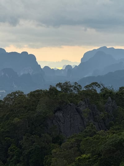 The image depicts a panoramic view of a mountainous landscape at dusk, with layers of mist obscuring the distant peaks. The foreground is dominated by a dense forest of lush green trees and dark, jagged rock formations. The middle ground features more forested hills, and the background consists of receding mountain ranges fading into the hazy horizon. A sliver of pale yellow sunlight is visible in the distance, peeking through a gap in the mountains, suggesting a sunset or sunrise. The sky above is a mosaic of muted gray and blue clouds. The location is identified as Ban Khao Tang, Thailand, a region known for its karst topography and tropical forests.