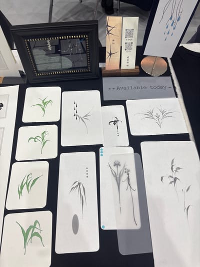 The scene depicts an overhead view of an art display table located in the city of Ban Rong Ruea Kham, Thailand. Various art pieces, primarily minimalist illustrations, are arranged on a dark, flat surface.
In the upper left, a black rectangular frame with gold bead accents encloses a reflective surface with a white and blue abstract design. To its right, two tall, narrow rectangular items, possibly bookmarks or art prints, stand upright in a small wooden holder. The left item features dark, stylized branch-like patterns and Thai script. The right item includes a QR code, similar branch patterns, and Thai script, with visible text "red ochre" and "อวยพร". A small, polished, circular metallic object rests to the right of these items.
Centrally positioned, a grey rectangular sign displays the text "--- Available today ---".
The majority of the table space is covered with white rectangular cards, each with rounded corners, featuring minimalist ink illustrations.
* The leftmost column contains four cards depicting green plant forms, possibly grasses or leaves.
* The center column displays three cards: the top shows a grey bamboo branch with three black teardrop shapes; the middle shows a black orchid-like flower with a single drip; the bottom shows grey bamboo leaves with four small black dots and a grey oval.
* The rightmost section includes two white cards with grey plant illustrations: one symmetrical, the other a multi-stemmed plant. Partially overlaying another white card in the bottom right is a translucent bookmark-like object featuring two slender, dark line-drawn flowers and two light blue dots on its upper left edge.
The background, blurred, reveals a segment of a person's lower legs and dark shoes, standing on a light-colored floor, suggesting an indoor setting.