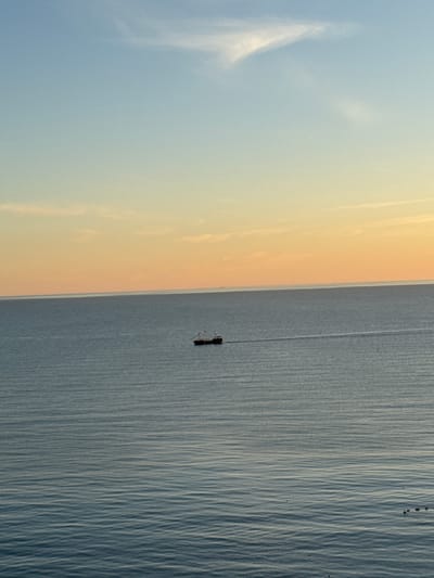 An aerial view captures a vast expanse of calm, deep blue water with subtle ripples under a gradient sky transitioning from pale blue at the top to a warm orange near the horizon. A solitary, dark-hulled vessel, appearing to be a small ship or a large boat with visible masts and rigging, is positioned in the mid-right section of the frame, creating a faint white wake behind it as it travels across the water. The overall scene conveys a sense of tranquility and open space, with no discernible human presence or urban elements visible within the frame. The location is identified as Varvarovka, Russia.