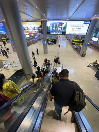 This is a photograph taken from an elevated perspective, looking down at a busy airport or large indoor transit terminal. The scene is brightly lit, suggesting daytime, though the interior lighting is prominent.

The primary subjects are numerous people, many of whom are carrying luggage, indicating travel. They are seen walking, standing, and riding on escalators. Some individuals are dressed casually, while others appear to be airport staff or security personnel. The setting is a modern, spacious terminal with a polished floor, large support columns, and numerous digital display screens and advertisements.

One prominent advertisement, visible in the upper left quadrant, is for "HONER HOMES," promoting "AN EXTRAORDINARY LIFE BEGINS HERE" with "Sprawling 5 BHK Mansions" in "Richmont" located at "Green Hills Road, Near HITEC City" with "POSSESSION BY DECEMBER 2026." Another advertisement, slightly lower and to the left, features an image of a futuristic building design and text including "THE STORY ABOVE 55 STOREYS" and a contact number. Overhead signs indicate directions to "Baggage Reclaim" and "Exit."

The activity taking place is the movement of travelers through the terminal. The presence of escalators suggests passengers are going to or from different levels of the building. The overall atmosphere appears to be one of organized transit, with people focused on their journeys. The orientation of the photograph is vertical, as if taken by someone holding their phone or camera at arm's length.