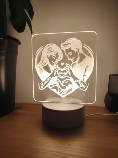 A close-up, eye-level shot features a personalized LED lamp with a heart design, illuminated from within. The engraving depicts a couple, with the woman on the left and the man on the right, both looking down at a baby in the center, also enclosed within the heart shape. The names "Mariana," "Melania," and "Petya" are etched into the design. The lamp sits on a light-colored, circular base. To the left, a dark pot with a large, dark green leaf is visible, partially obscuring the background. To the right, a black electronic device is situated, with a white power cord extending from the lamp's base and leading towards it. The objects are placed on a wooden surface with a visible grain. The background is a textured, light-colored wall. The lighting suggests an indoor environment, likely a home or office. The location is not determinable from the image.