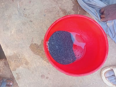 A red plastic bowl sits on a rough, light-colored surface with brown stains and dry grass. Inside the bowl, dark-colored sand or granules fill about half the space with some water present. To the left, there's a glimpse of a gray cloth and a dark-skinned hand. A white sandal-clad foot with dusty toes is visible in the upper left corner, and another foot wearing sandals can be seen in the upper right, positioned on a ledge-like surface.