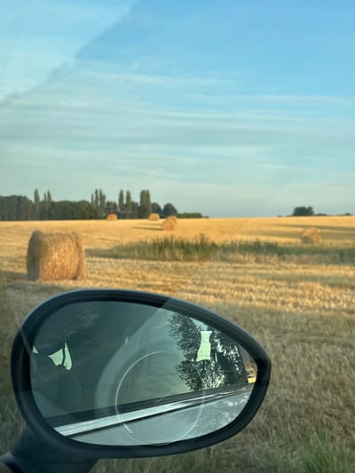 The image depicts a rural scene with hay bales scattered across a harvested field under a blue sky with wispy clouds. In the foreground, a car's side mirror reflects the passing landscape, including trees and the interior of the car, with a silhouette of a person visible. The scene appears to be located in the countryside near Trouville-Alliquerville, France, given the characteristic flat, open fields of the Pays d'Auge region. The lighting suggests it is either early morning or late afternoon, with the sun casting a warm glow on the dry hay. No specific actions or interactions are evident, other than the implied movement of the car capturing the reflection.