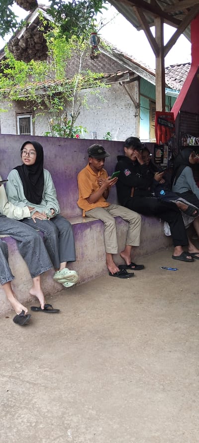Four people are sitting on a concrete bench with a purple surface in front of a store in Cimaung, Indonesia, located at the coordinates -7.074 latitude and 107.547 longitude.  The person on the far left is wearing a gray hijab, a green jacket, and dark jeans. The person on the far right is wearing a gray hijab and a blue jacket. The person in the middle is wearing a yellow shirt and khaki pants. The person on the right is wearing a black jacket and black pants. They are all looking at their phones and appear to be in their late teens or early twenties. There is a tree in the background. The scene is a casual gathering of friends.  The store is in a residential area.  The ground is concrete and there are several items on the floor, including sandals.  The people appear to be relaxed and comfortable. There is no obvious interaction between them other than a shared space.  It appears to be a warm, sunny day.  There is no activity occurring in the background.  The scene is quiet and calm.