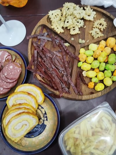 On a table, a platter of assorted snacks is arranged. The platter is divided into sections, one holding numerous thin, dark red strips of jerky. Another section contains small, brightly colored puffed snacks in shades of yellow, green, and orange. A third section holds a pile of light-colored, irregularly shaped crackers with a snowflake-like pattern.

To the left of the platter, a blue and yellow patterned plate displays several sliced portions of a rolled food item, appearing to be a roulade with alternating layers of yellow and white. Adjacent to this plate, another blue and yellow patterned plate holds several thick, circular slices of a reddish-pink sausage with visible fat marbling.

In the lower right corner of the image, a clear plastic container holds a pile of thinly sliced, light yellow snacks, possibly potato sticks. A white wine glass with a broad base is partially visible in the upper left corner. The overall scene suggests a casual gathering or a snack spread. The lighting is even, indicating it could be indoors during daylight. No text is visible in the image. The setting appears to be a domestic environment.