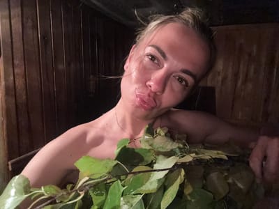 A woman with blonde hair pulled up is posing for a selfie in a wooden sauna. She is partially submerged in what appears to be a wooden tub or pool, with a bundle of wet green leaves resting on her chest and shoulder. Her lips are puckered. The walls of the sauna are constructed of dark vertical wooden planks. The lighting is dim, suggesting the interior of a sauna.